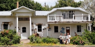 House with 3 men in front and with a dog and a cat next to them. The house has a big sign that says "FOR SALE"